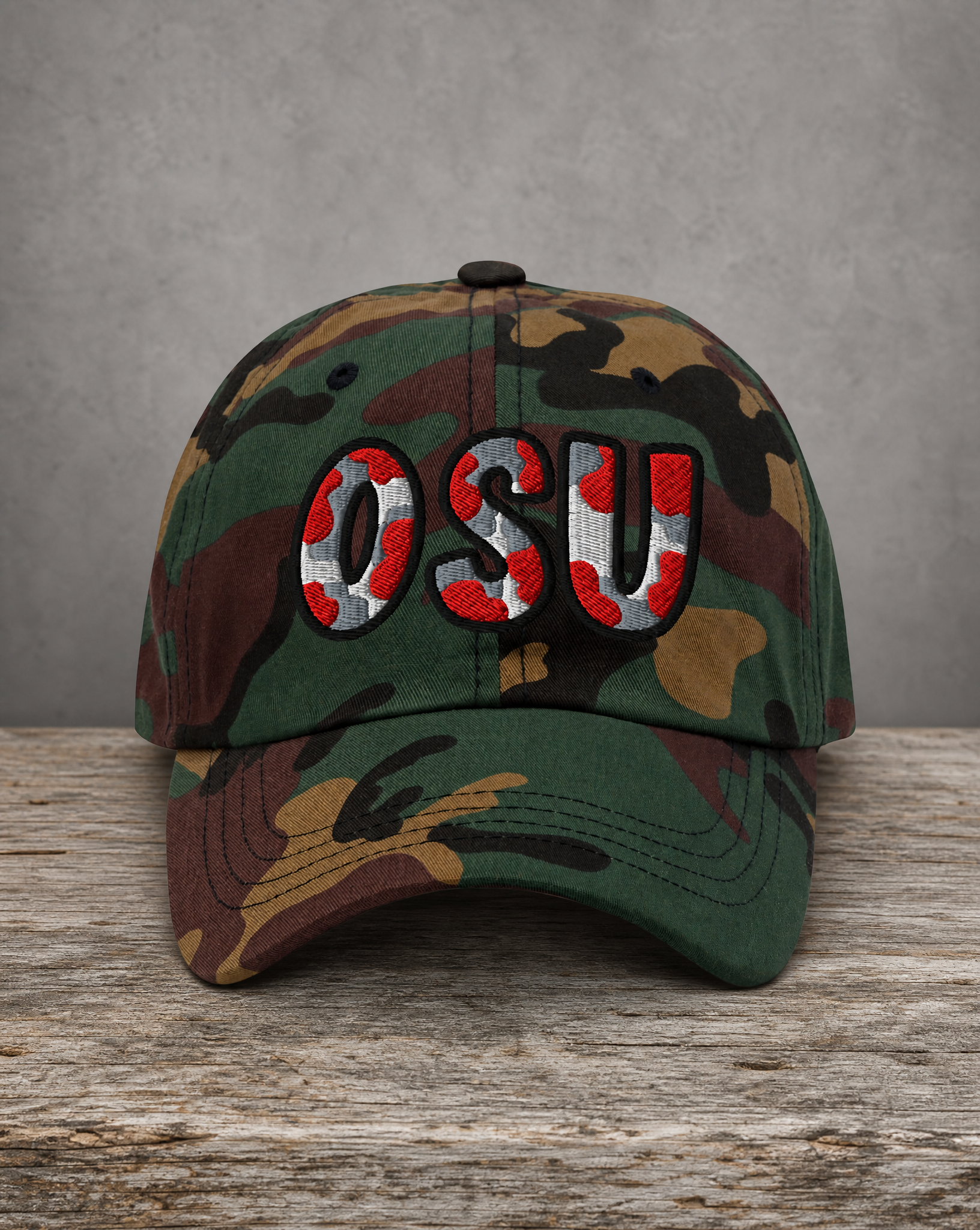 A camouflage baseball cap with 'OSU' embroidered on the front in red, white, and black, resting on a rustic wooden surface against a gray textured background.
