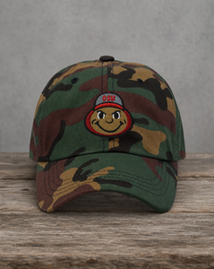 A camouflage baseball cap resting on a rustic wooden surface against a textured gray wall. The cap features a green, brown, and tan camo pattern with a central embroidered patch depicting a smiling cartoon face wearing a gray cap with 'OSU' embroidered in red.