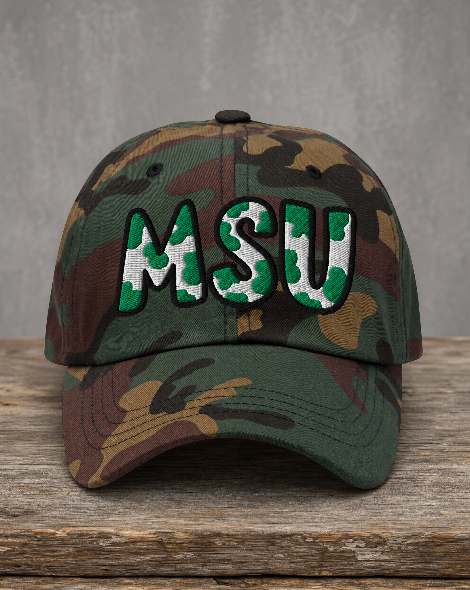 A camouflage baseball cap with 'MSU' embroidered on the front in green, white, and black, resting on a rustic wooden surface against a gray background.