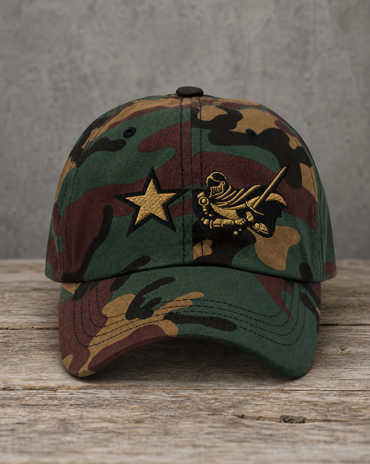 A camouflage baseball cap resting on a rustic wooden surface against a textured gray background. The cap features a green, brown, and tan woodland camo pattern with a gold embroidered star and a gold knight emblem on the front.