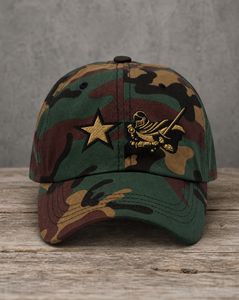 A camouflage baseball cap resting on a rustic wooden surface against a textured gray background. The cap features a green, brown, and tan woodland camo pattern with a gold embroidered star and a gold knight emblem on the front.