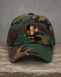 A camouflage baseball cap with a cartoon mascot embroidered on the front, displayed on a rustic wooden surface against a textured gray background.