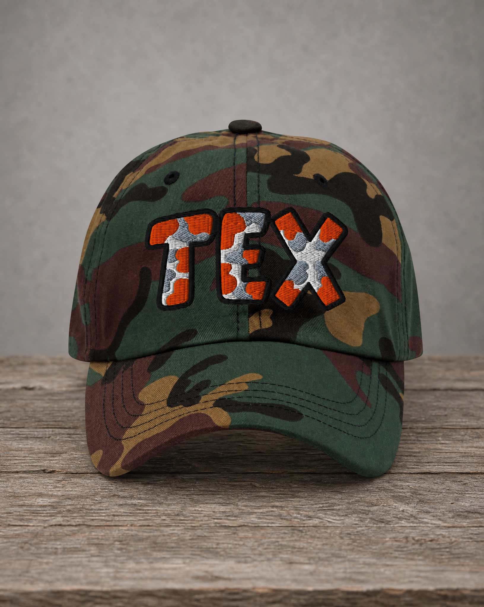 A camouflage baseball cap with 'TEX' embroidered on the front in orange and silver, resting on a rustic wooden surface against a neutral gray background.