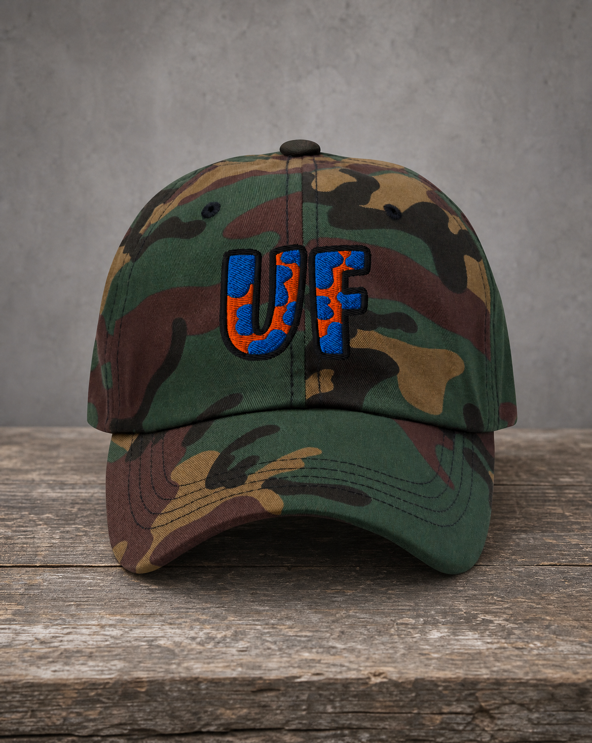 A camouflage baseball cap with embroidered 'UF' lettering in blue and orange, resting on a rustic wooden surface against a textured gray background.