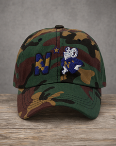 A camouflage baseball cap with embroidered navy blue and gold letter 'N' and a white and blue mascot patch on the front, displayed on a rustic wooden surface against a gray background.