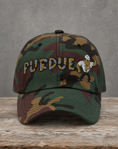 A camouflage baseball cap with 'PURDUE' embroidered in gold and blue, featuring a small Purdue mascot patch on the side, displayed on a rustic wooden surface against a gray background.