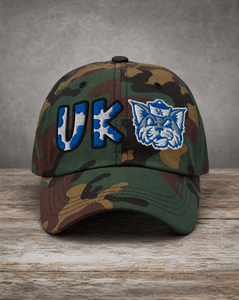 A camouflage baseball cap resting on a rustic wooden surface against a textured gray background. The cap features embroidered blue and white 'UK' lettering and a blue wildcat mascot logo on the front panels.