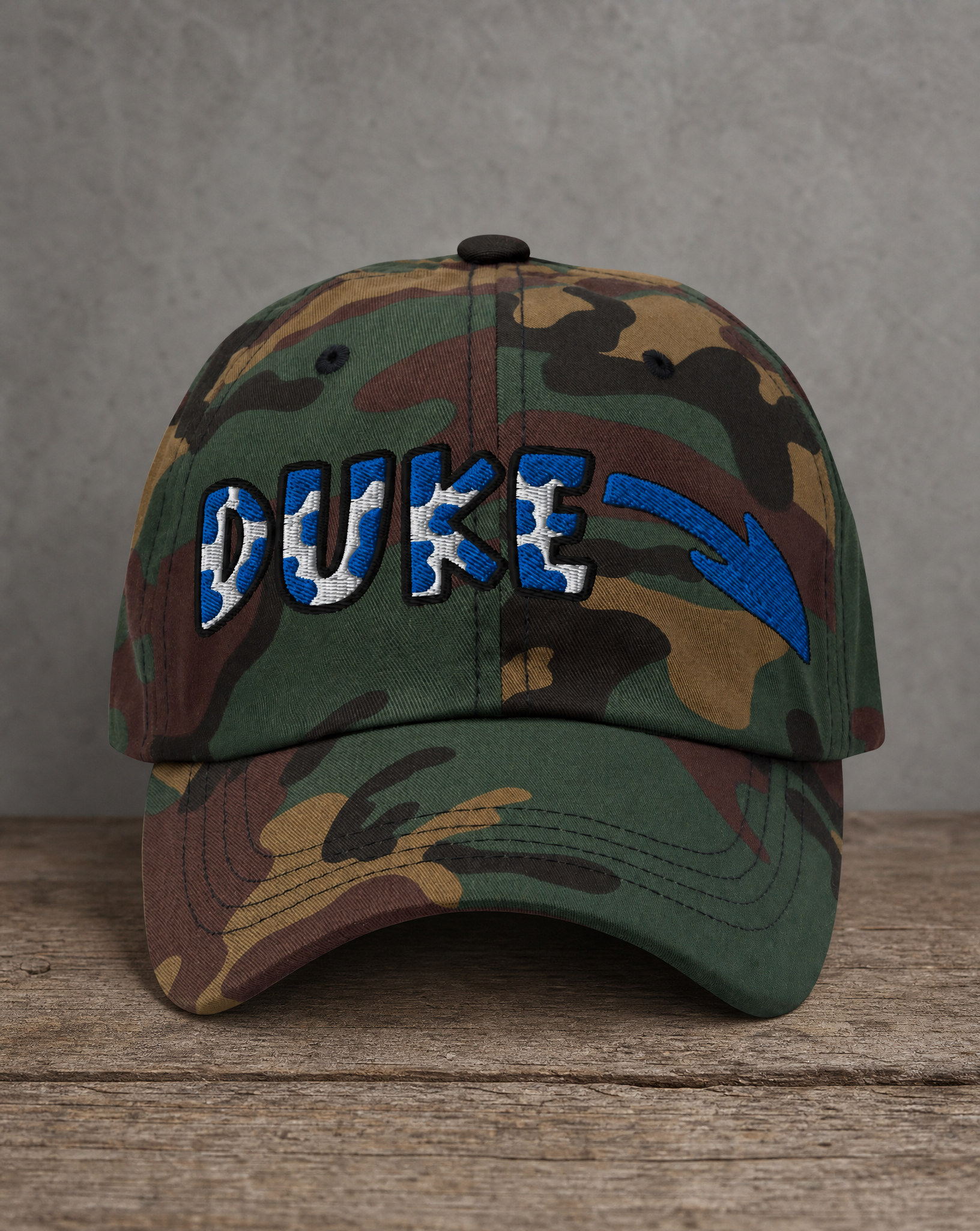 A camouflage baseball cap with 'DUKE' embroidered in blue and white with a blue arrow, resting on a rustic wooden surface against a gray background.