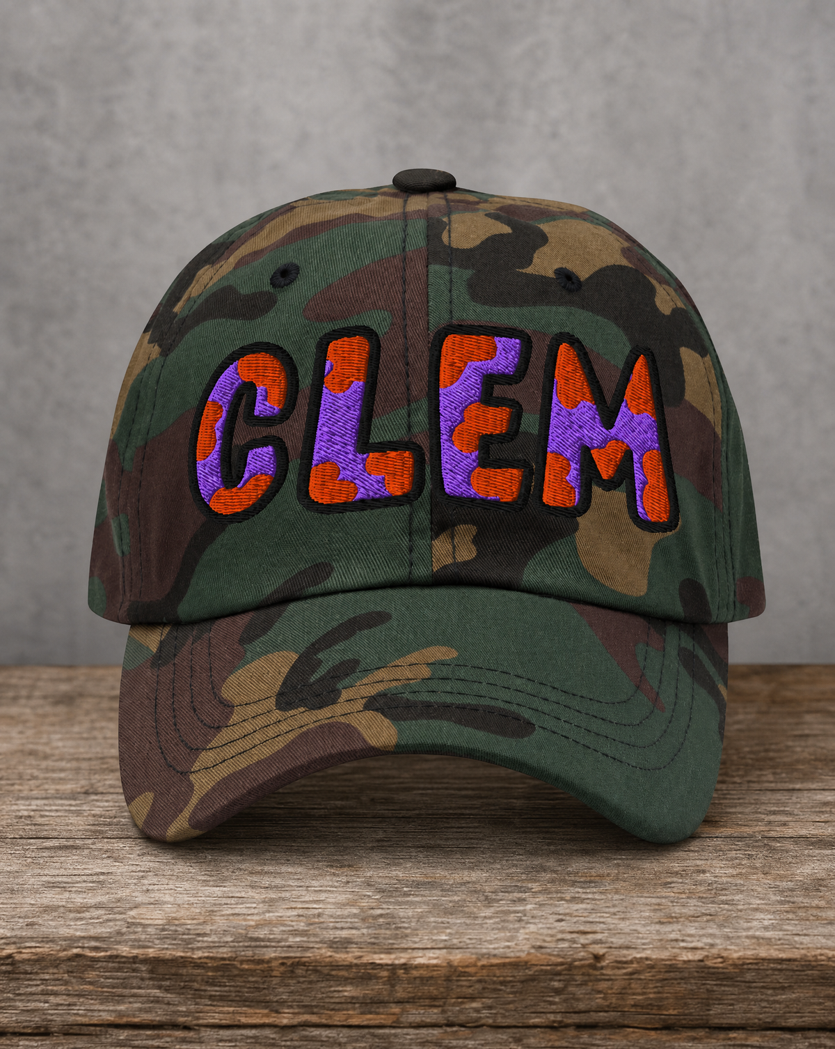A camouflage baseball cap with 'CLEM' embroidered in orange and purple on the front, displayed on a rustic wooden surface against a textured gray wall.