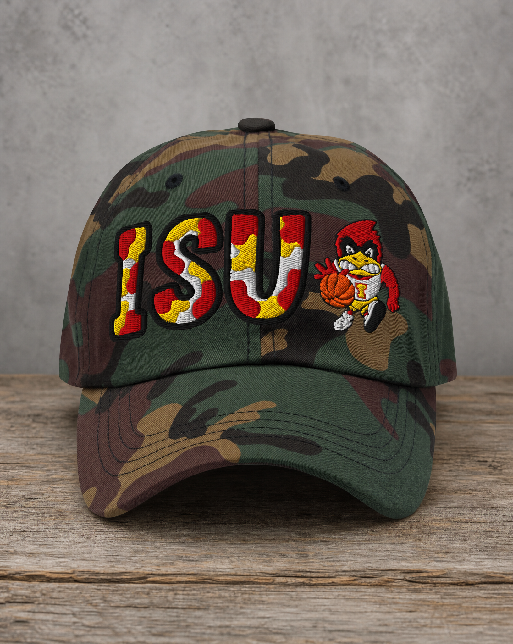 A camouflage baseball cap featuring a large embroidered 'ISU' logo in red, yellow, and white, alongside a cartoon mascot holding a basketball, displayed on a rustic wooden surface against a gray concrete wall.