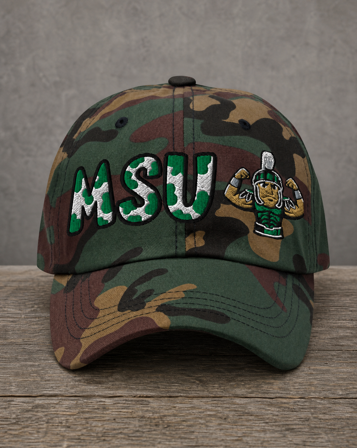 A camouflage baseball cap featuring 'MSU' embroidered in green and white with a mascot character on the front, displayed on a rustic wooden surface against a gray background.