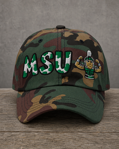 A camouflage baseball cap featuring 'MSU' embroidered in green and white with a mascot character on the front, displayed on a rustic wooden surface against a gray background.