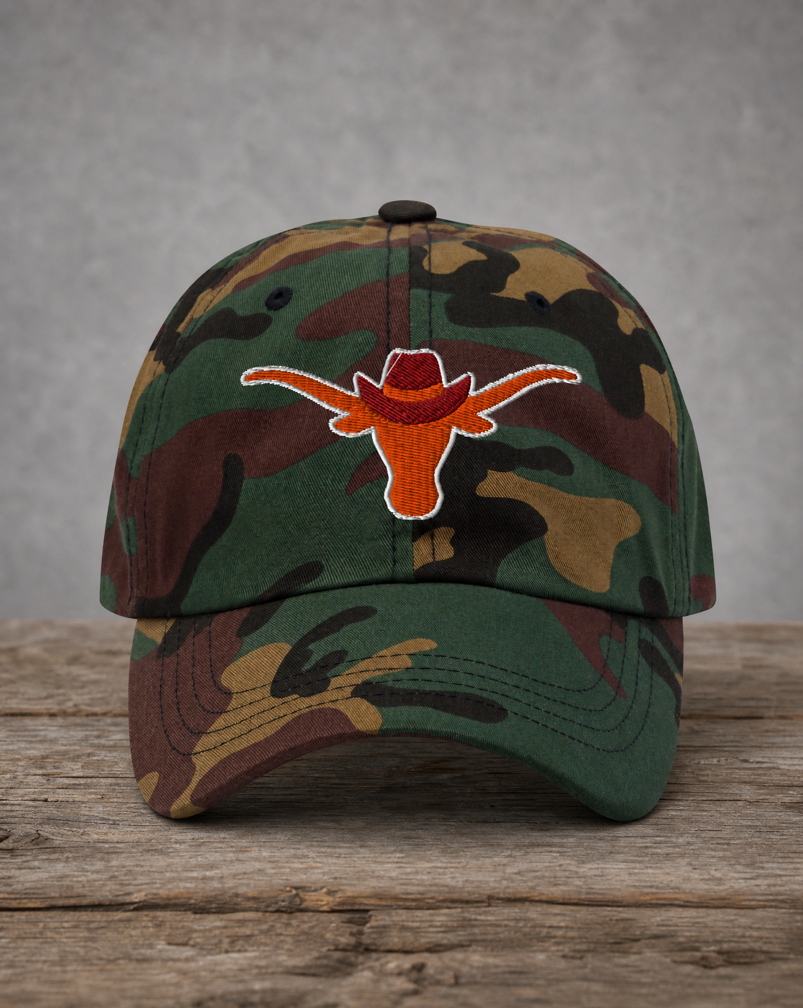 A camouflage baseball cap featuring an embroidered orange Longhorn cowboy hat logo on the front, displayed on a rustic wooden surface against a gray background.