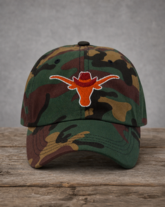 A camouflage baseball cap featuring an embroidered orange Longhorn cowboy hat logo on the front, displayed on a rustic wooden surface against a gray background.