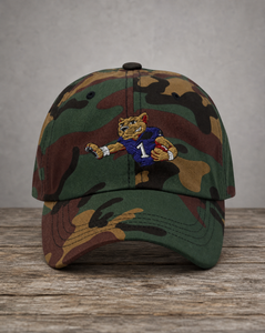 A camouflage baseball cap featuring an embroidered wildcat mascot wearing a purple football jersey with the number 1, displayed on a rustic wooden surface against a neutral gray background.