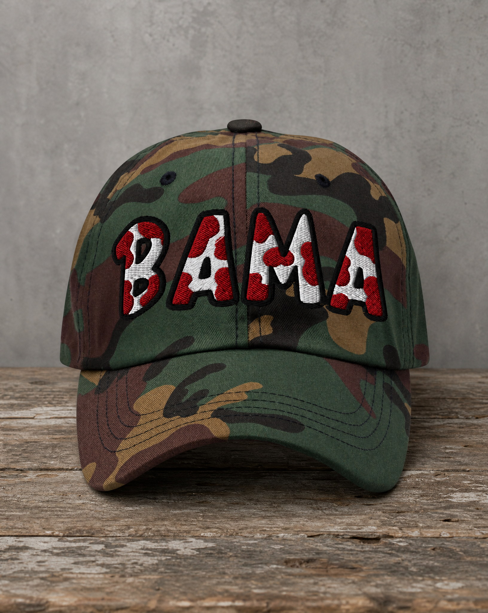 A camouflage baseball cap with 'BAMA' embroidered across the front in red, white, and black, resting on a rustic wooden surface against a gray concrete wall.