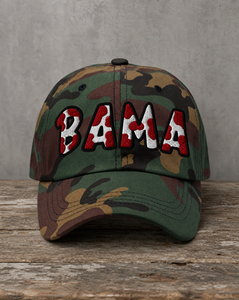 A camouflage baseball cap with 'BAMA' embroidered across the front in red, white, and black, resting on a rustic wooden surface against a gray concrete wall.