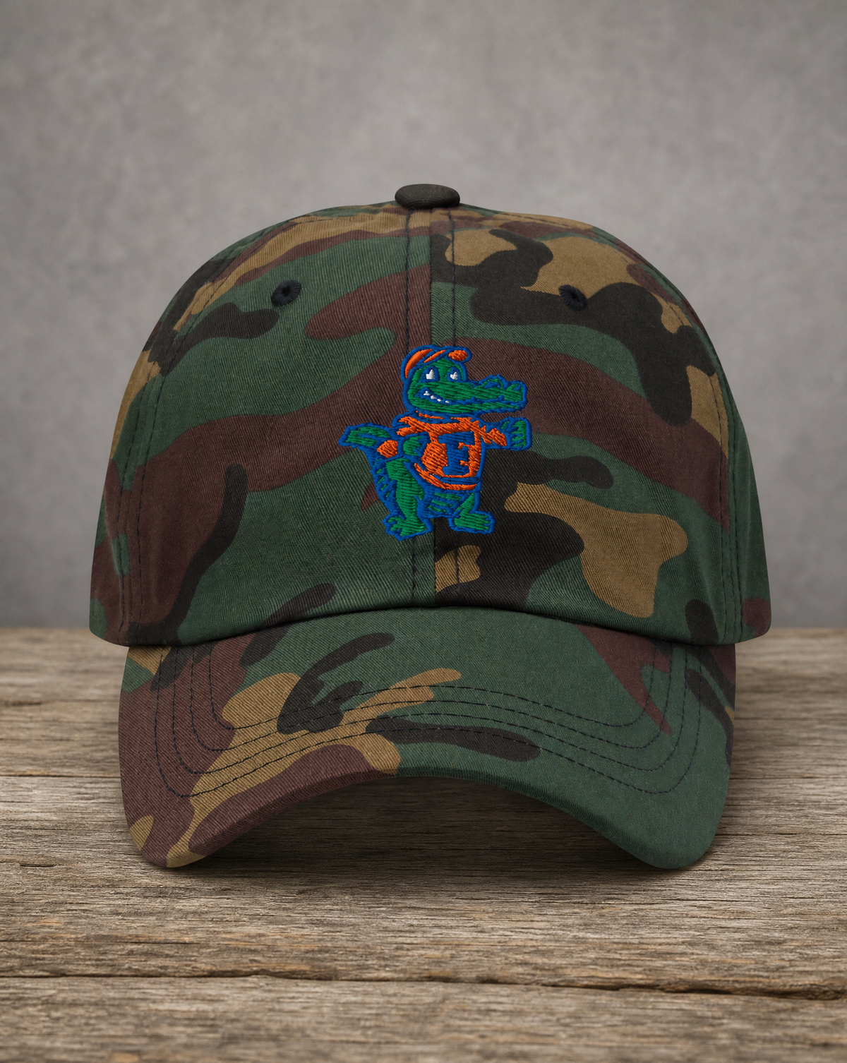 A camouflage baseball cap resting on a rustic wooden surface against a textured gray background. The cap features a green, brown, and tan woodland camo pattern with a central embroidered alligator mascot wearing a blue and orange shell.