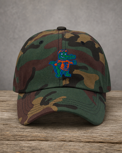 A camouflage baseball cap resting on a rustic wooden surface against a textured gray background. The cap features a green, brown, and tan woodland camo pattern with a central embroidered alligator mascot wearing a blue and orange shell.