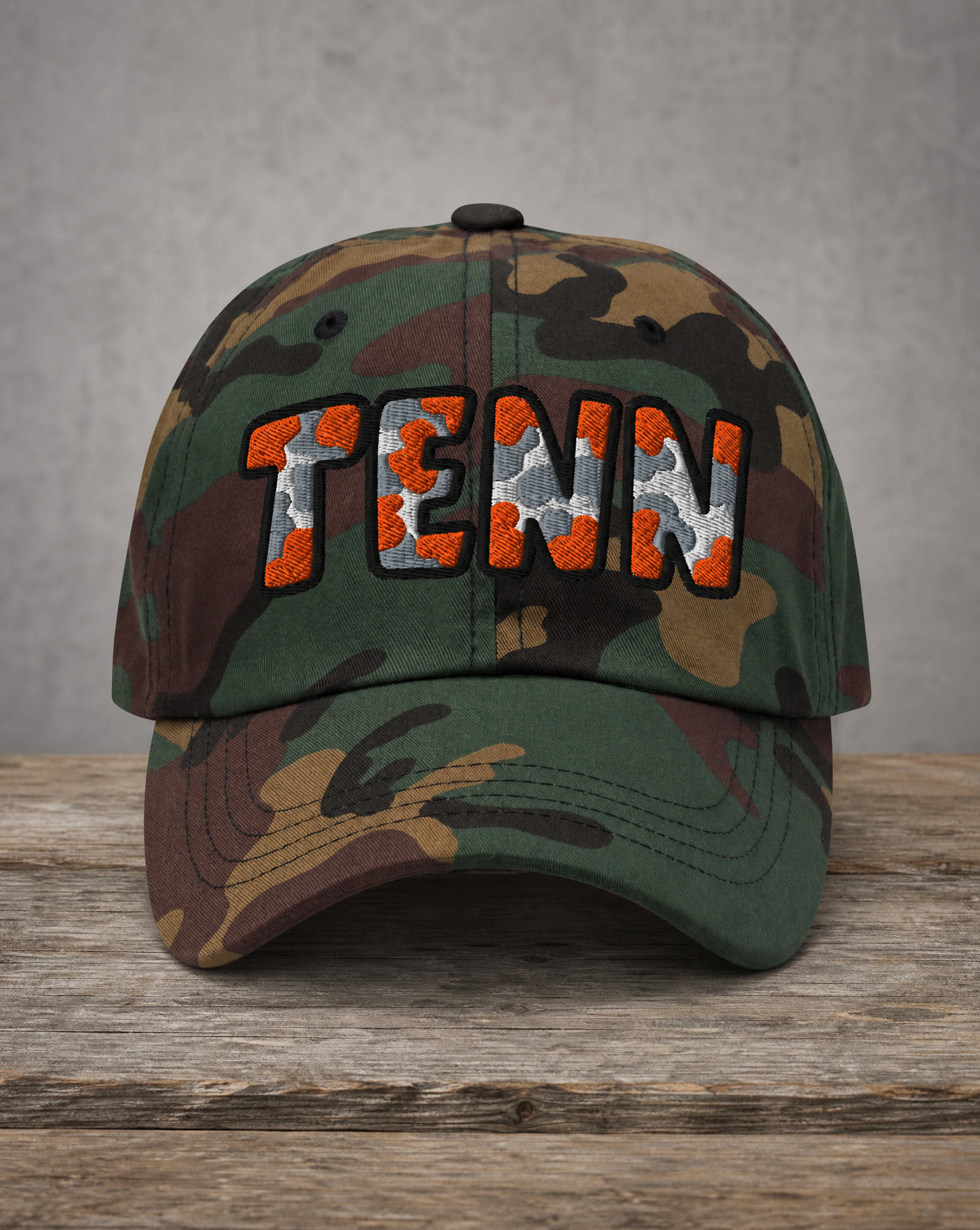 A camouflage baseball cap with 'TENN' embroidered on the front in orange, white, and gray, resting on a rustic wooden surface against a textured gray background.