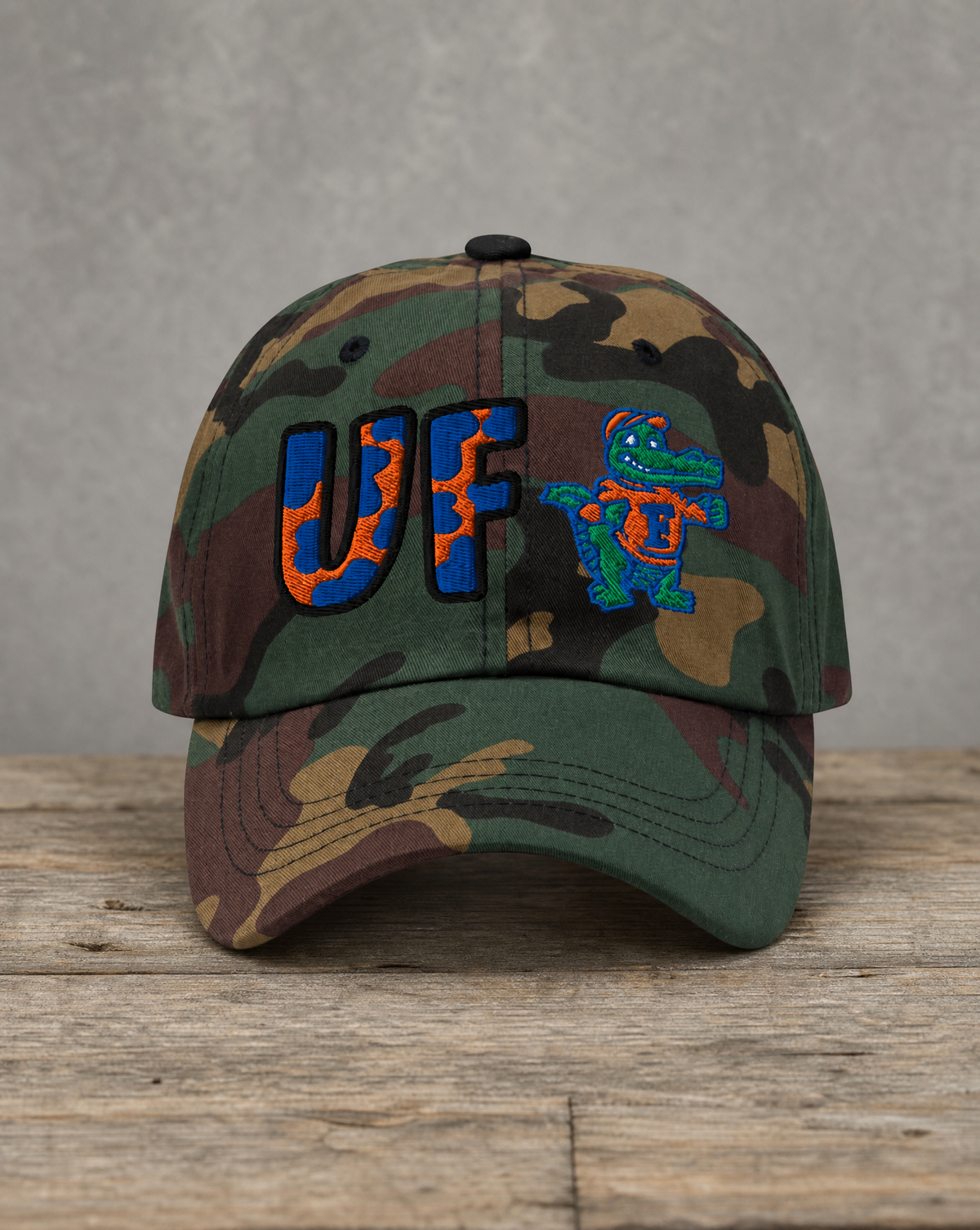 A camouflage baseball cap featuring embroidered 'UF' lettering in blue and orange with a Florida gator mascot patch, displayed on a rustic wooden surface against a gray background.