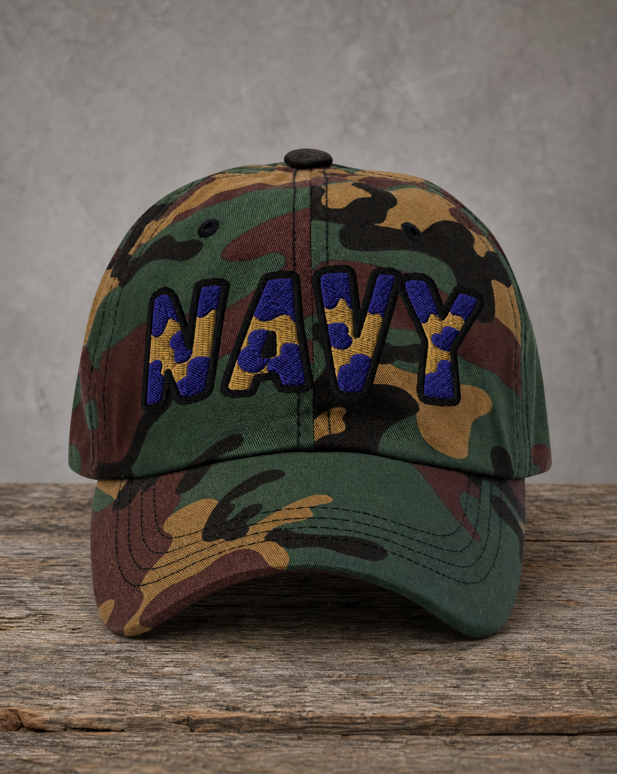 A camouflage baseball cap with 'NAVY' embroidered in blue and yellow on the front, resting on a rustic wooden surface against a textured gray wall.