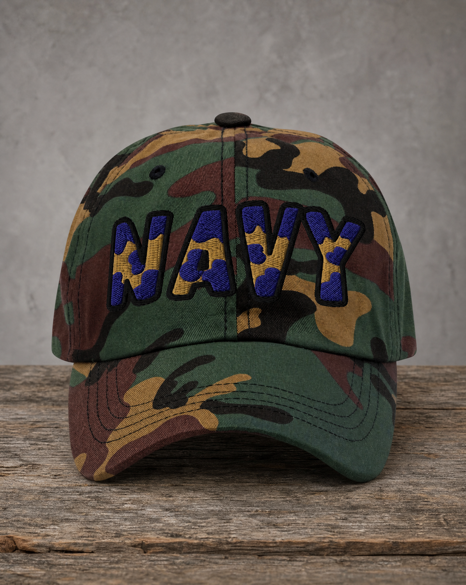 A camouflage baseball cap with 'NAVY' embroidered in blue and yellow on the front, resting on a rustic wooden surface against a textured gray wall.
