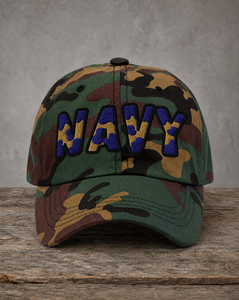 A camouflage baseball cap with 'NAVY' embroidered in blue and yellow on the front, resting on a rustic wooden surface against a textured gray wall.