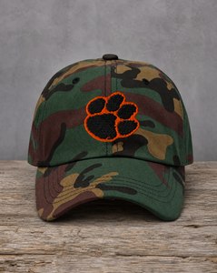 A camouflage baseball cap with a black paw print patch outlined in orange on the front, displayed on a rustic wooden surface against a textured gray background.