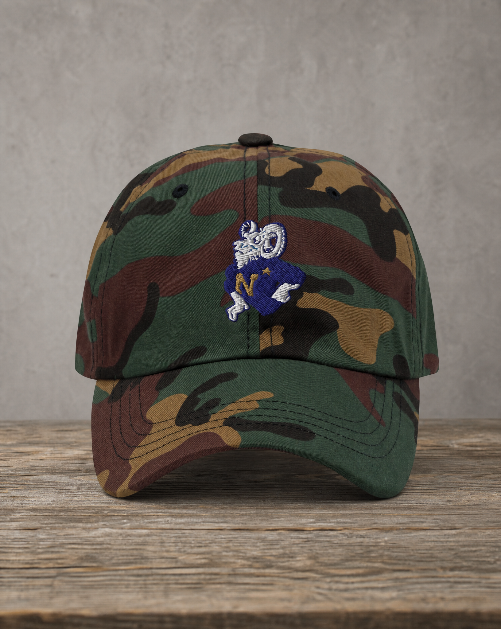 A camouflage baseball cap with a small embroidered bulldog mascot on the front, displayed on a rustic wooden surface against a textured gray background.