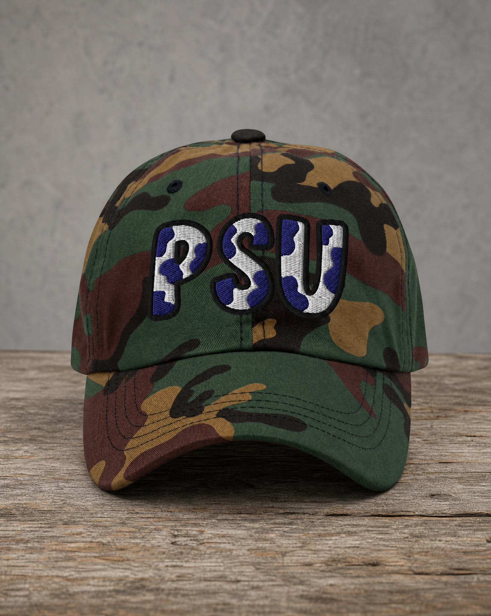 A camouflage baseball cap with 'PSU' embroidered in blue and white on the front, resting on a rustic wooden surface against a gray textured background.