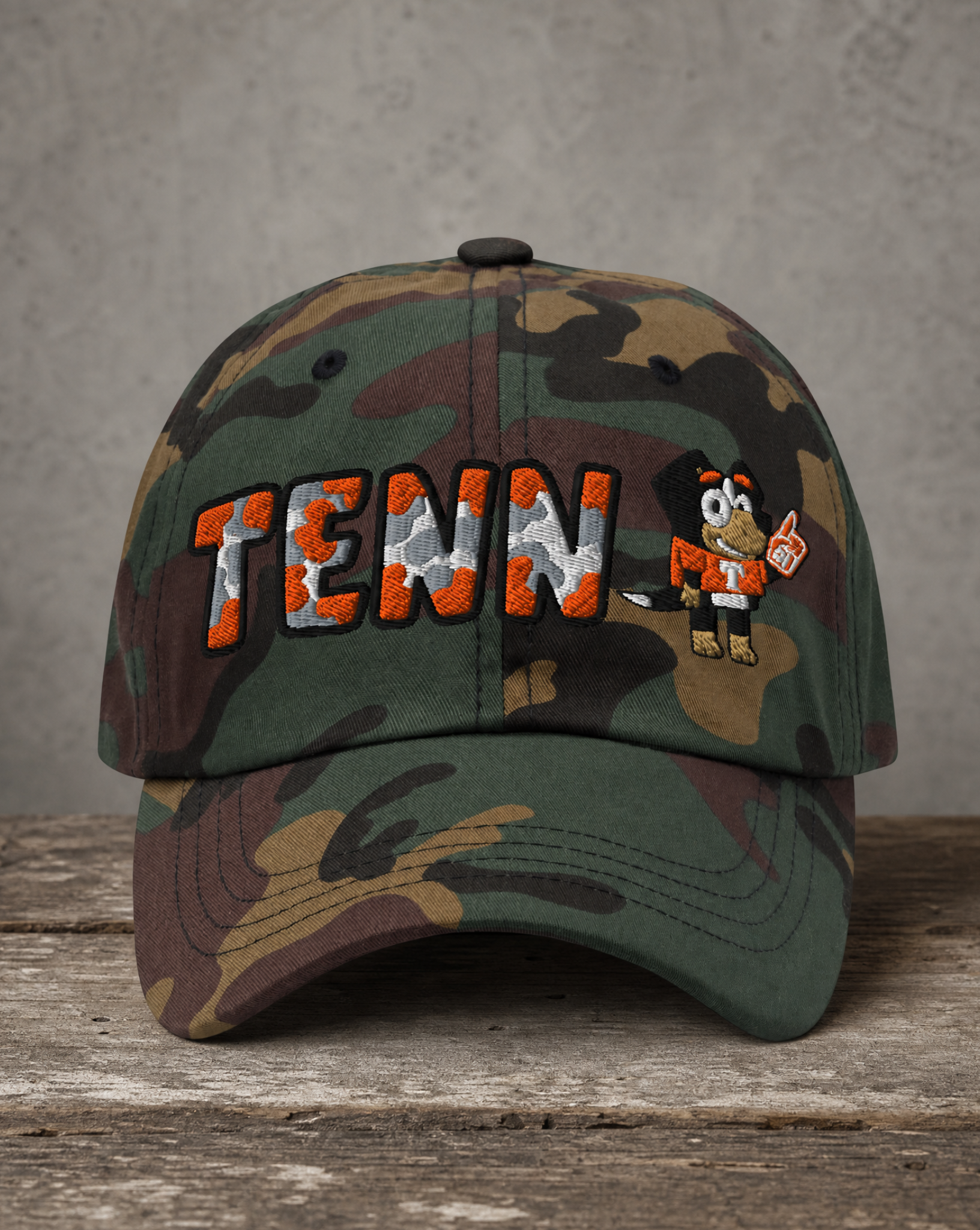A camouflage baseball cap resting on a rustic wooden surface against a textured gray wall. The front of the cap features 'TENN' embroidered in orange and white camouflage lettering with black outlines, alongside a cartoon mascot wearing an orange and white uniform.