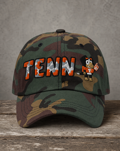 A camouflage baseball cap resting on a rustic wooden surface against a textured gray wall. The front of the cap features 'TENN' embroidered in orange and white camouflage lettering with black outlines, alongside a cartoon mascot wearing an orange and white uniform.