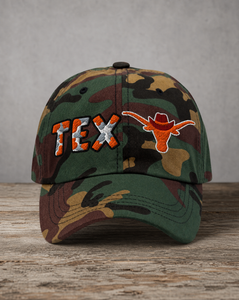 A camouflage baseball cap featuring 'TEX' and a Longhorn logo embroidered on the front, resting on a rustic wooden surface against a neutral gray background.