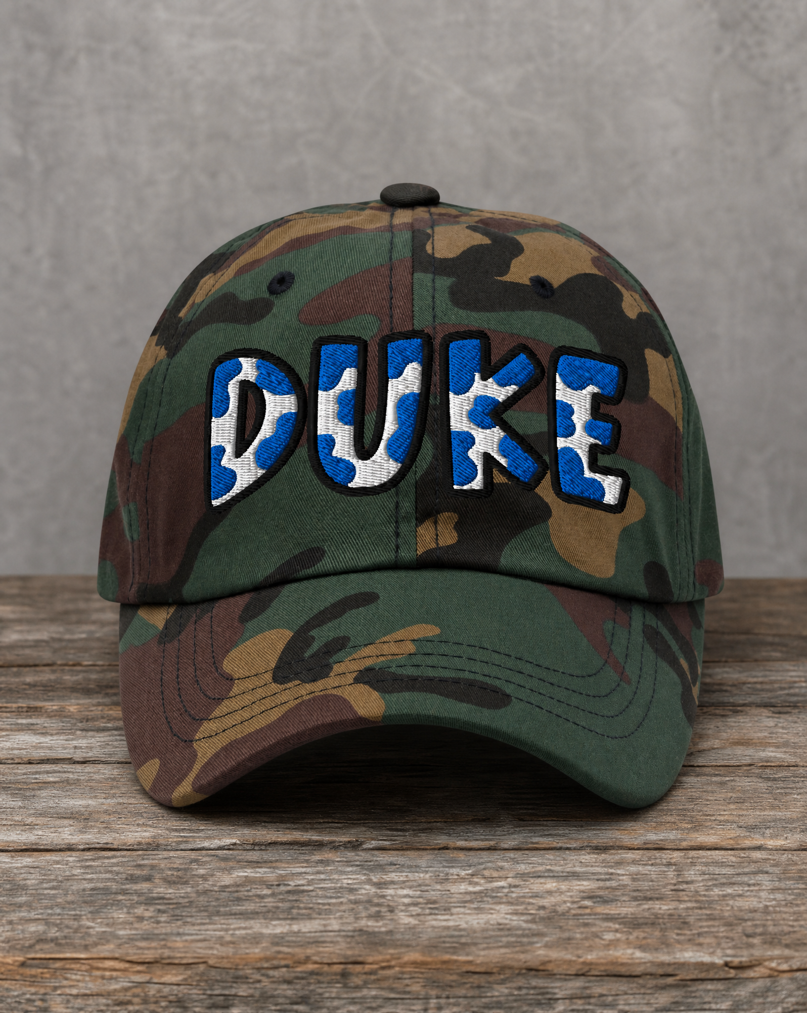 A camouflage baseball cap with 'DUKE' embroidered across the front in blue and white, resting on a rustic wooden surface against a gray textured background.