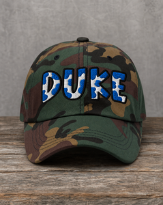 A camouflage baseball cap with 'DUKE' embroidered across the front in blue and white, resting on a rustic wooden surface against a gray textured background.
