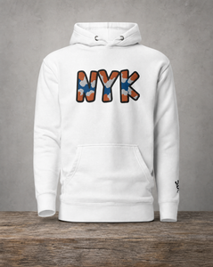A white pullover hoodie displayed on a wooden surface against a textured gray wall. The front of the hoodie features a large graphic spelling 'NYK' in a camouflage pattern of orange, blue, and white, outlined in black.