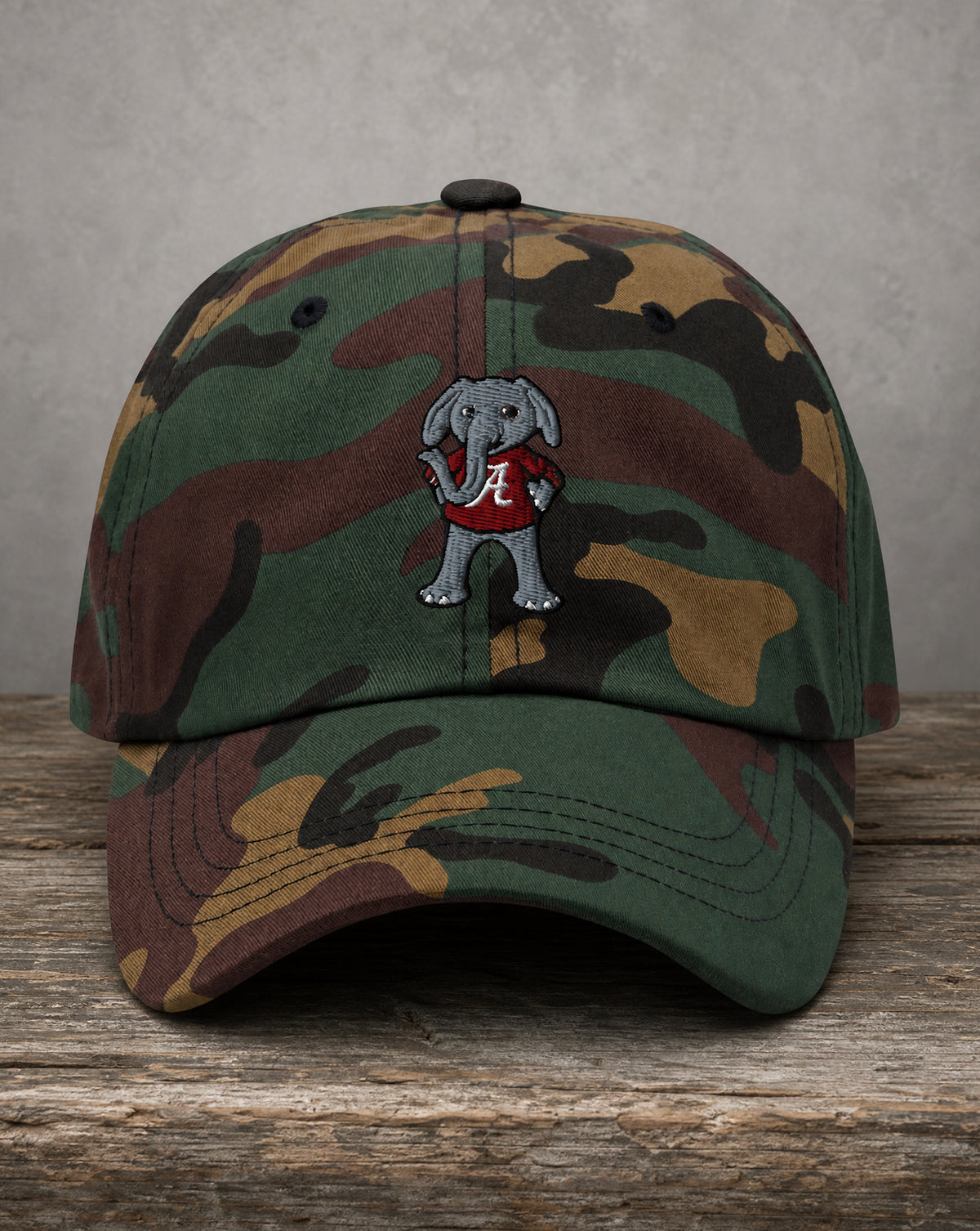 A camouflage baseball cap featuring an embroidered gray elephant mascot wearing a red shirt with a white 'A' logo, displayed on a rustic wooden surface against a textured gray background.