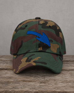 A camouflage baseball cap with a prominent blue arrow embroidered on the front, resting on a rustic wooden surface against a neutral gray background.