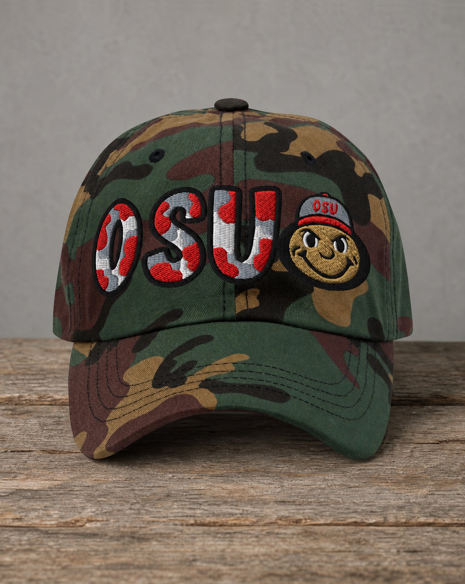 A camouflage baseball cap with 'OSU' embroidered in red, white, and blue on the front, alongside a smiling mascot face wearing an OSU cap. The cap is displayed on a rustic wooden surface against a neutral gray background.
