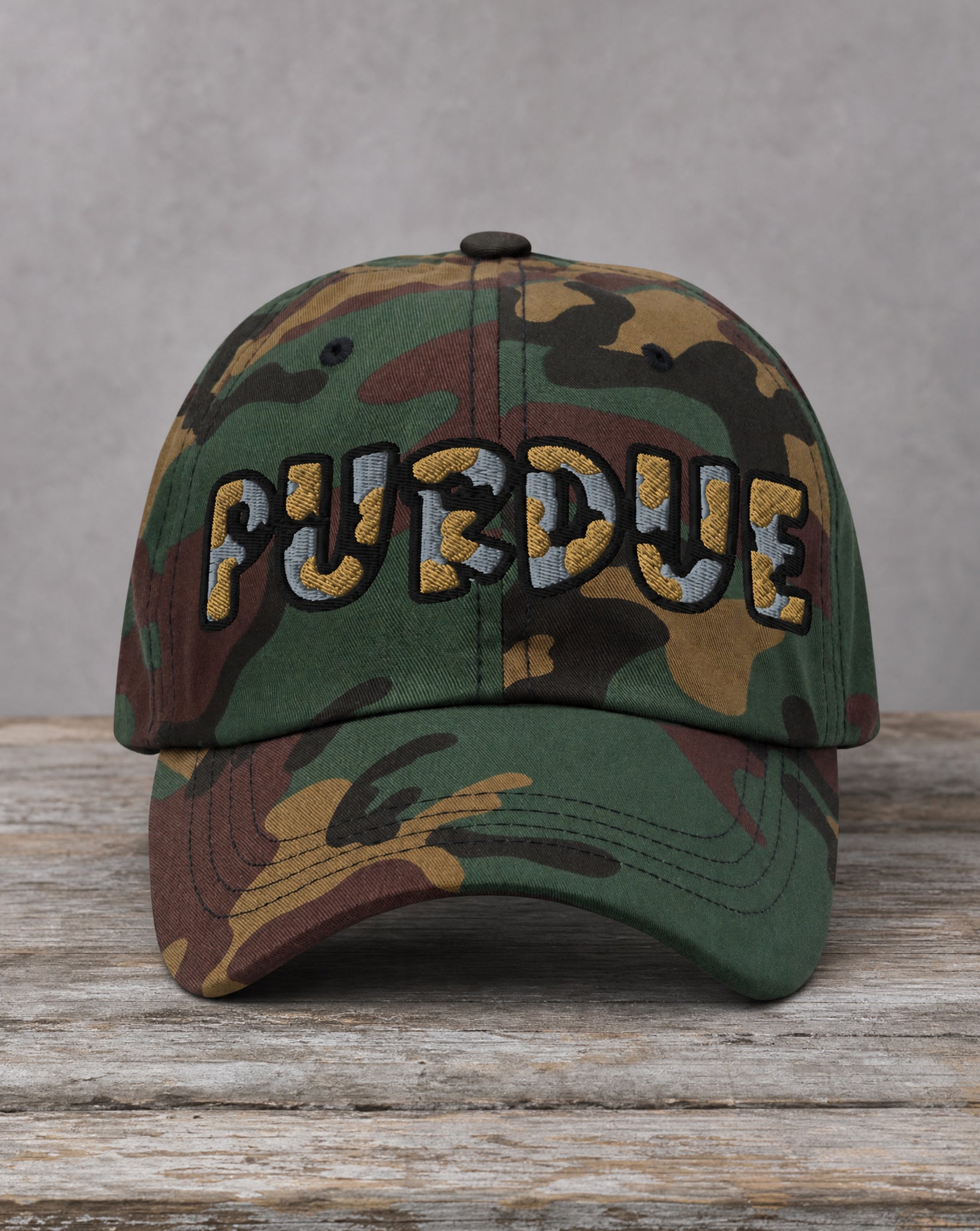 A camouflage baseball cap with 'PURDUE' embroidered across the front in a gold and gray patterned font, resting on a rustic wooden surface against a gray background.