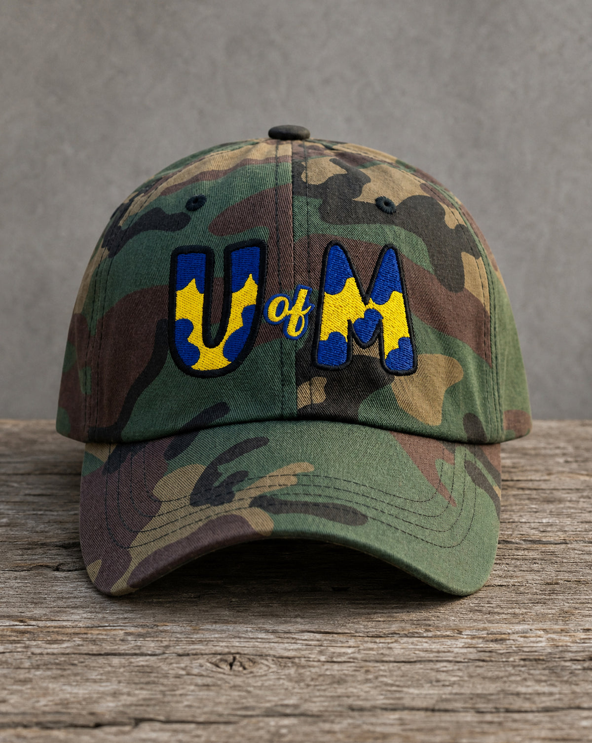 A camouflage baseball cap resting on a rustic wooden surface against a gray background. The front features embroidered letters spelling 'UM' in blue and yellow, with 'of' stitched in yellow script between them.