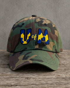 A camouflage baseball cap resting on a rustic wooden surface against a gray background. The front features embroidered letters spelling 'UM' in blue and yellow, with 'of' stitched in yellow script between them.